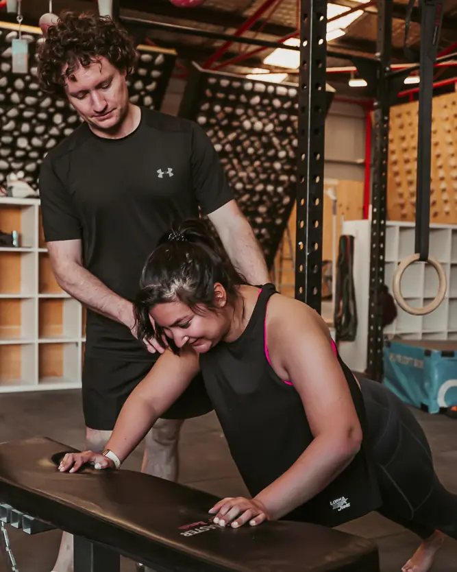 Rick standing behind a client, guiding her through an incline push-up on a bench